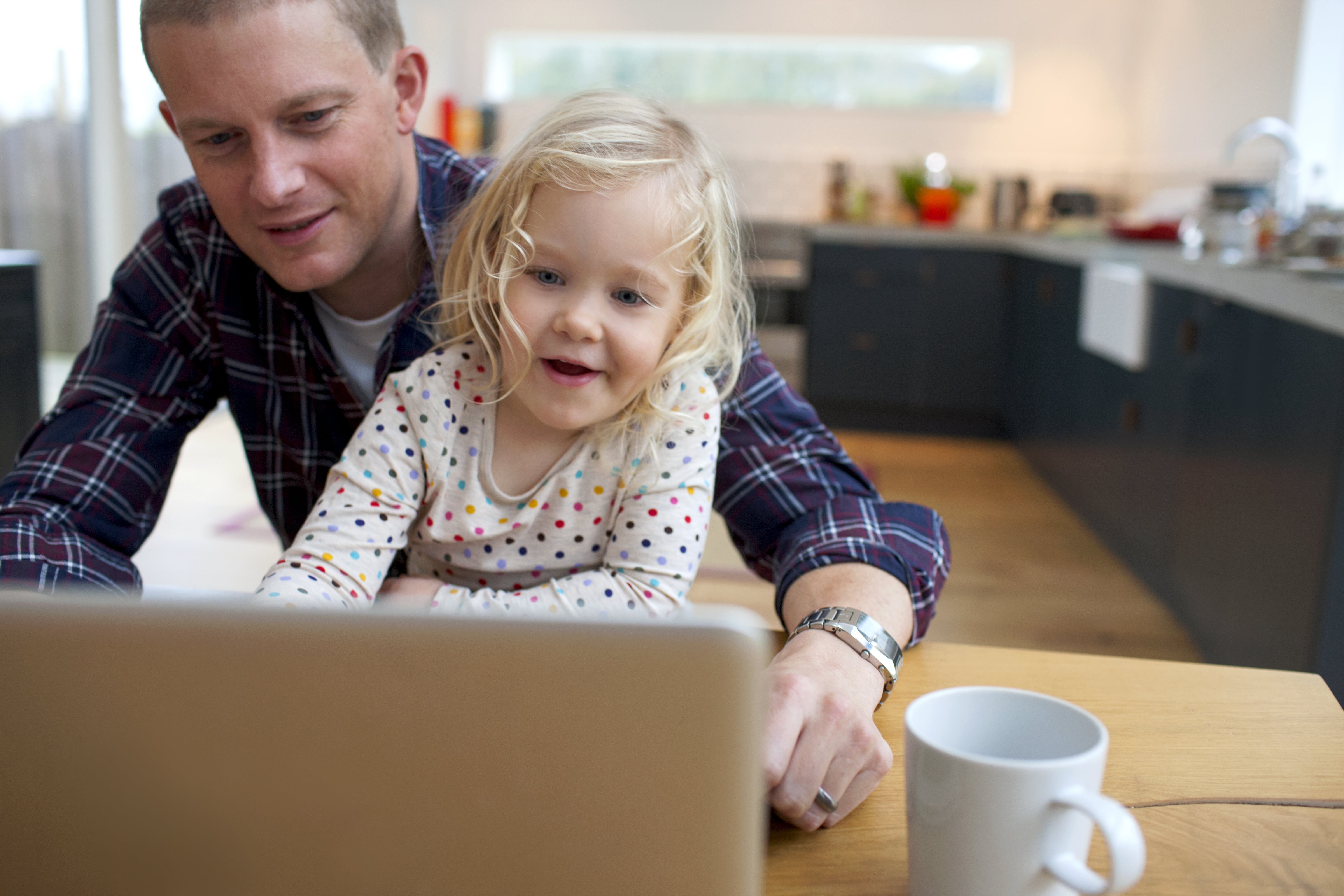 Person med rutete skjorte og klokke på håndleddet, sitter ved bord med kaffekopp og bærbar PC.