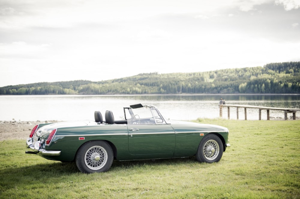 Grønn veteranbil parkert på en gresslette ved en innsjø, med skog i bakgrunnen.